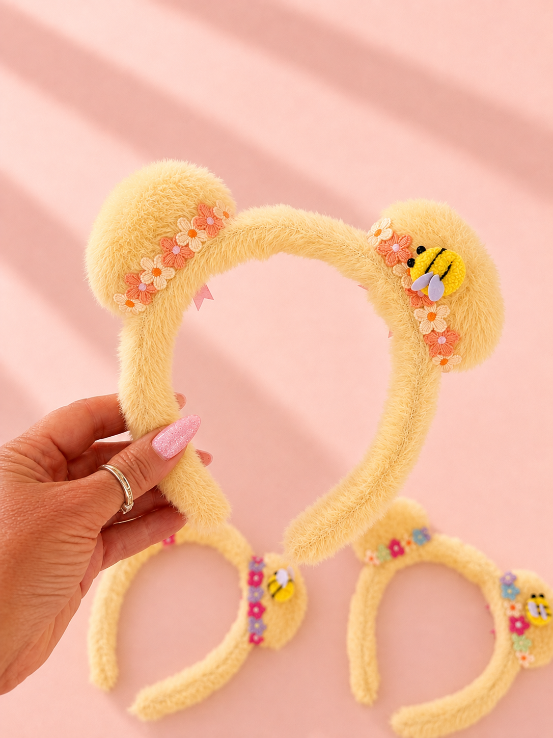 A hand holds a fuzzy pale yellow Mickey ear headband decorated with small flowers and a bee applique, with two matching headbands laid on a soft pink background.