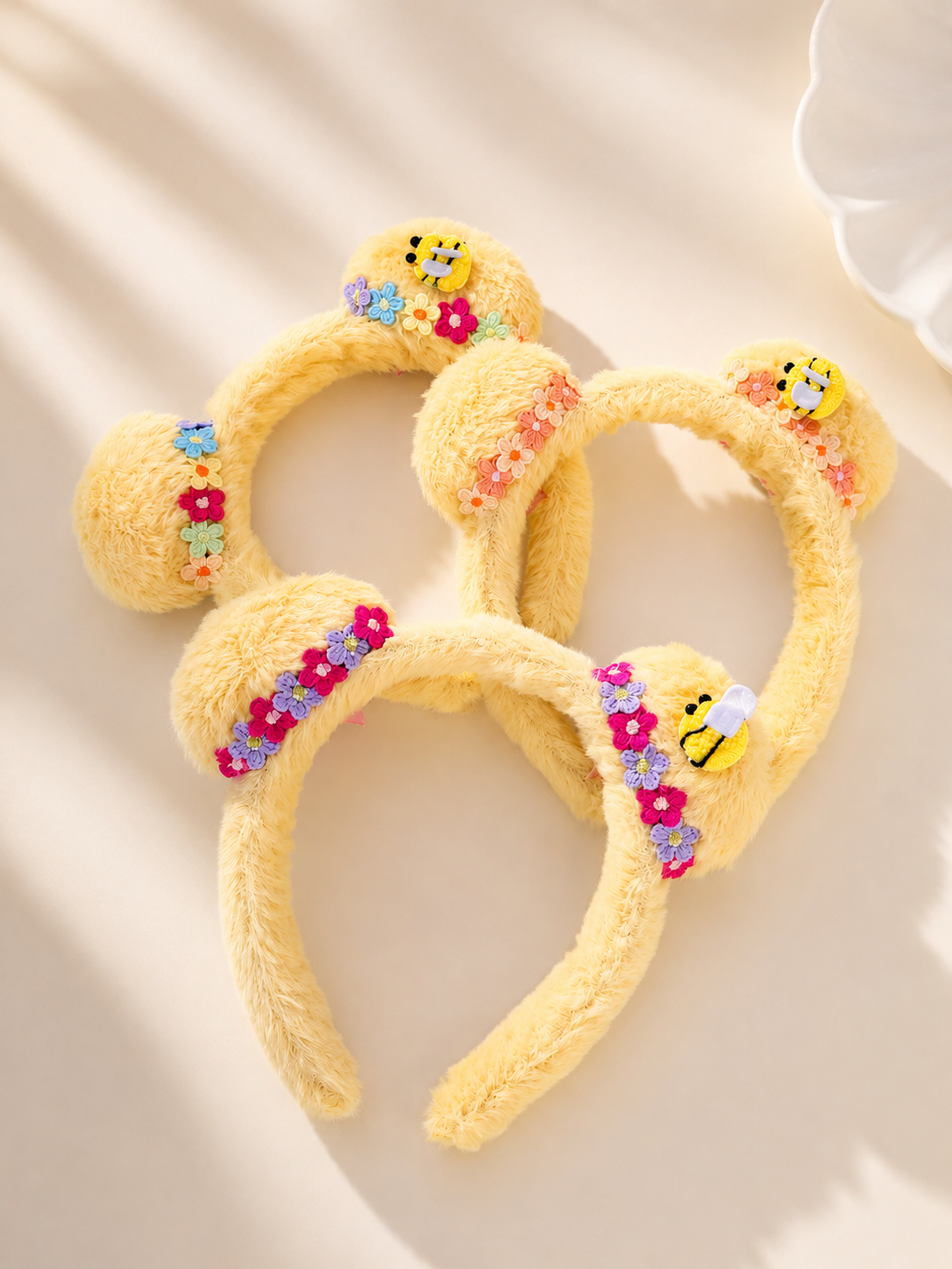 Three yellow fuzzy Mickey ear headbands decorated with colorful sequins, flowers, and small bee embellishments, arranged on a warm beige surface with soft shadows.