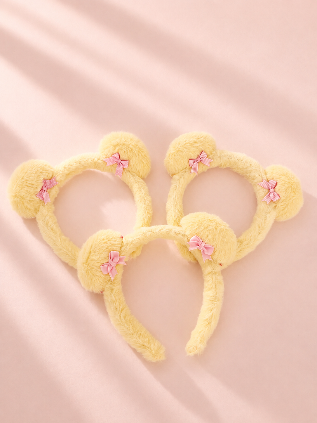 Three yellow fuzzy Mickey mouse ear headbands with small pink bows, arranged on a soft pink background with gentle shadows.