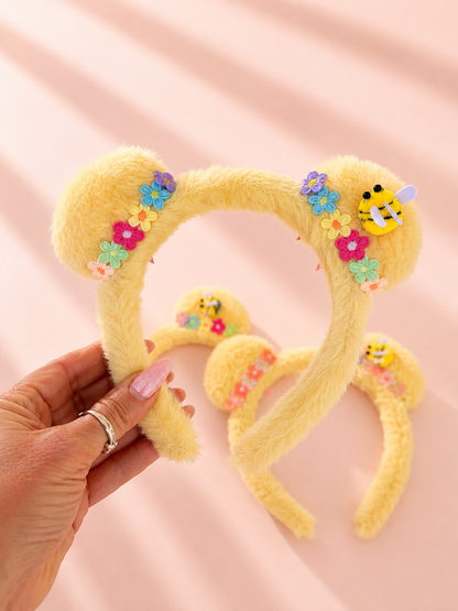 A yellow fuzzy plush mickey ear headband decorated with colorful flowers and a bee applique, held by a hand against a soft pink background with sun rays.