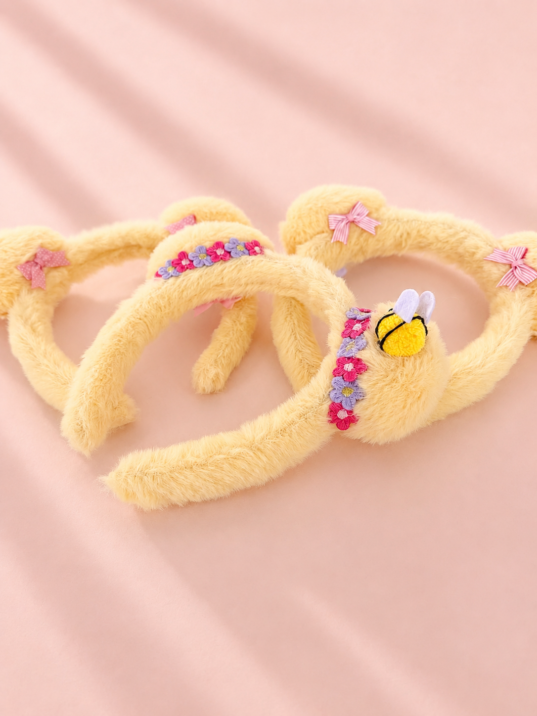 Three yellow fuzzy Mickey mouse ear headbands arranged on a soft pink fabric background. One headband features a small embroidered bee on a floral band, and two others have pink bow accents.