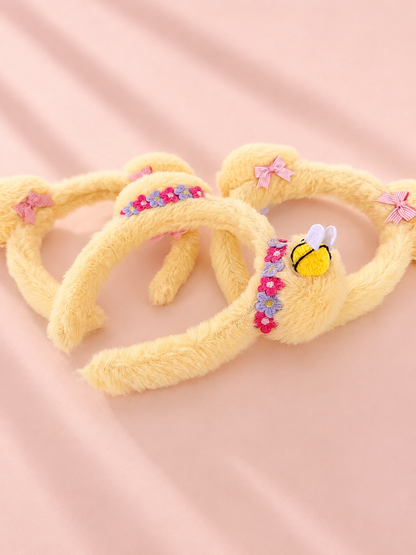 Three yellow fuzzy Mickey mouse ear headbands arranged on a soft pink fabric background. One headband features a small embroidered bee on a floral band, and two others have pink bow accents.