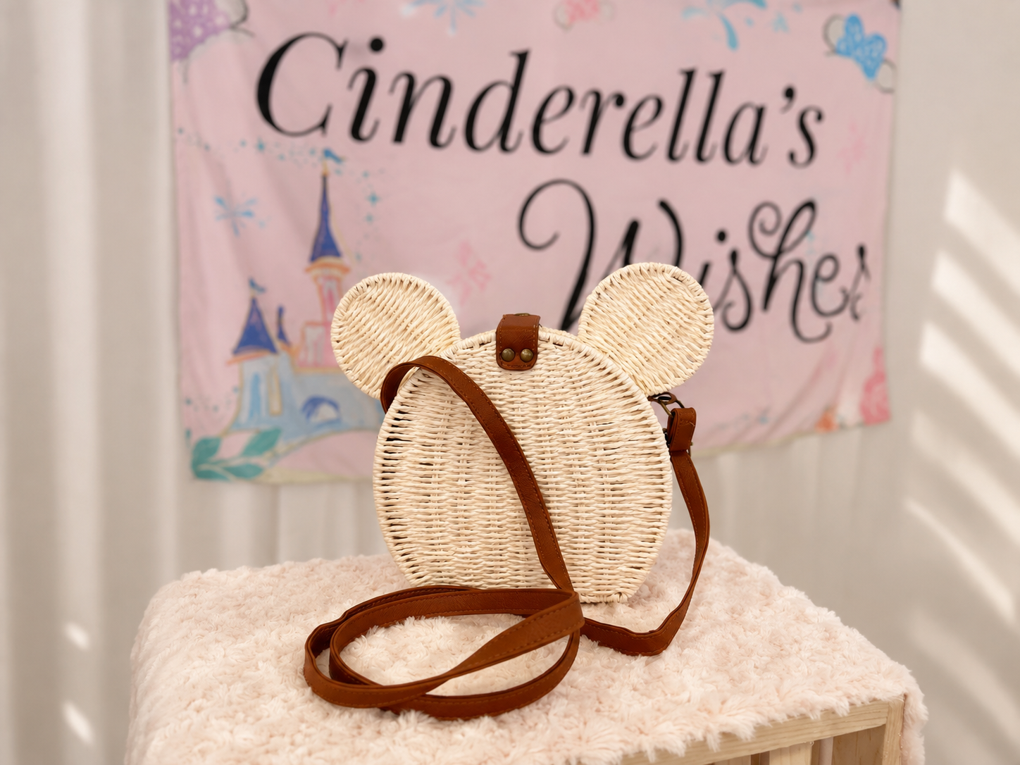 A woven straw crossbody bag shaped like a mouse head with round ears and a brown strap, displayed on a fluffy cream surface in front of a pink 'Cinderella's Wisher' banner.