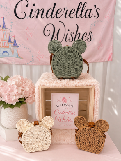 Three woven mouse-ear backpacks in cream, sage green, and natural tan are displayed on a pink table runner in front of a 'Cinderella's Wishes' banner. A plush pink box with a 'Welcome' sign stands in the center, flanked by pink flowers on the left.