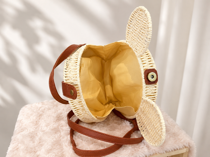 A woven straw Mickey -shaped shoulder bag with long floppy ears, a mustard yellow interior lining, and brown faux-leather straps, displayed on a pink faux-fur surface against a softly lit white curtain background.